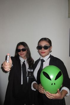 two women dressed in business attire and sunglasses holding an alien balloon while giving the thumbs up