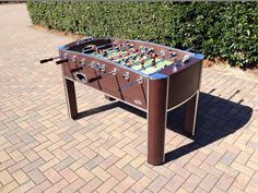 a foosball table sitting on top of a brick sidewalk next to a bush