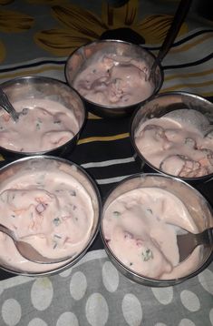 four silver bowls filled with food on top of a tablecloth covered floor next to two spoons