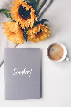 an open notebook with the word sunday written on it next to a cup of coffee and sunflowers