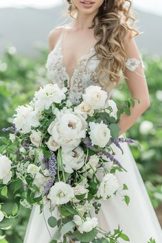 a woman in a wedding dress holding a bouquet of flowers and greenery with her hands behind her back