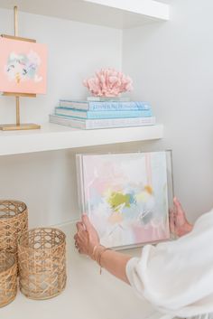 a woman holding up a painting in front of some baskets on a white shelf next to other items