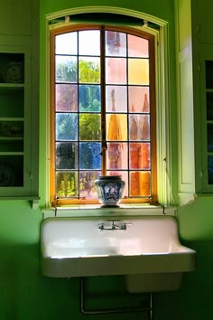 a white sink sitting under a window next to a green wall and cupboards with bottles on it