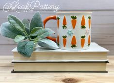 an orange and white coffee mug sitting on top of a book next to a plant