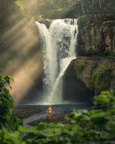 a woman standing in front of a waterfall with sunlight coming through the leaves on it