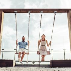 a man and woman sitting on swings in front of a wooden frame, smiling at the camera