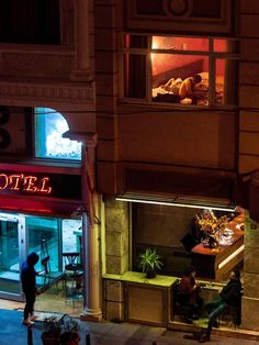 two people sitting on benches in front of a hotel at night with the lights on