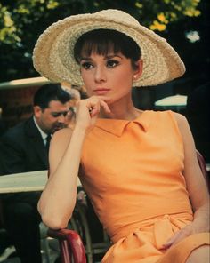 a woman in an orange dress and straw hat sitting on a chair with her hand under her chin