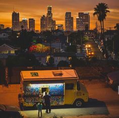 two people standing in front of a food truck at night with the city skyline behind them