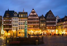 many buildings are lit up at night in an old european town with fountain and clock tower