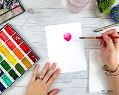 a person is holding a paintbrush and painting on paper with watercolors next to it