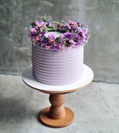 a purple cake with flowers on top sitting on a wooden stand in front of a gray wall