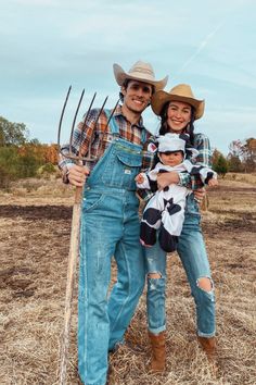 a man and woman in overalls holding a baby while standing on dry grass with pitchforks