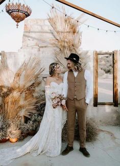 a bride and groom standing next to each other in front of a wall with dried plants