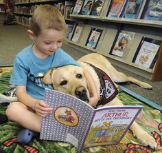 a young boy reading a book to a dog on the floor in front of bookshelves