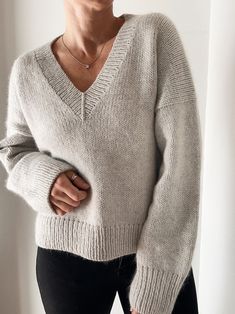 a woman wearing a gray sweater and black pants standing in front of a white wall