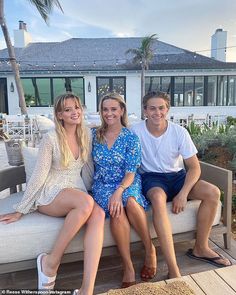 three people are sitting on a bench in front of a house and one person is smiling at the camera