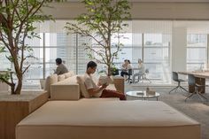 two men are sitting on couches in an office setting with large windows and potted trees