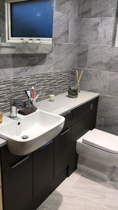 a white toilet sitting next to a bathroom sink under a window in a gray tiled wall