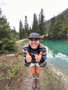 a woman giving the thumbs up while standing in front of a lake with mountains and trees