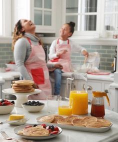 two women in the kitchen with breakfast foods on the counter and pancakes, orange juice, and muffins