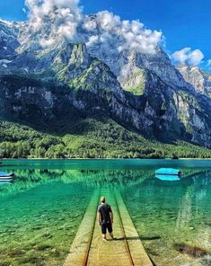 a man walking across a wooden bridge over water with mountains in the background and blue sky