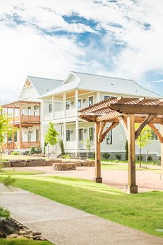 a wooden gazebo sitting in the middle of a lush green field next to a large white house