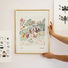 a person holding up a framed painting next to other drawings and artwork on the wall