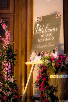 a welcome sign sitting on top of a wooden table next to pink and purple flowers