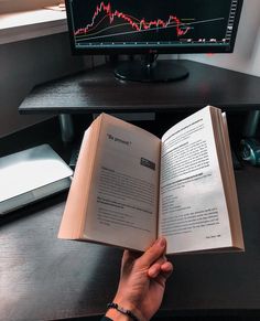 a person is holding an open book in front of a computer screen with a stock chart on it