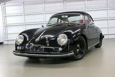 an old black car parked in a garage next to a white wall and two doors