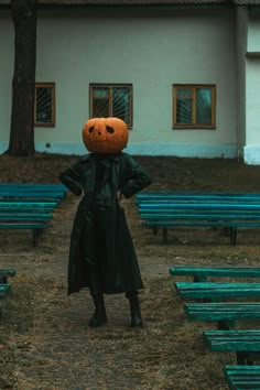 a person in a black coat with a pumpkin head on their head standing next to green benches