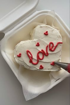 a heart shaped cake with the word love spelled in red icing on it, sitting in a styrofoam container