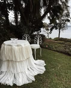 two white chairs and a table in the grass near some trees with water in the background