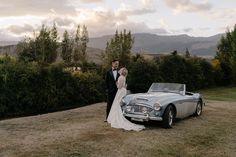 a bride and groom standing in front of an old car on the side of a hill