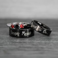 two black rings with red stones are sitting on a wooden table, one has the words simply mean written on it