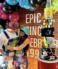 a man and woman standing next to each other in front of a brick wall with balloons