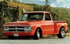 an orange pickup truck parked in front of a forest with mountains and trees behind it