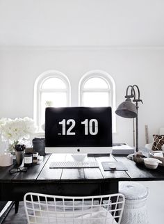 a desk with a computer on it in front of two large windows and white flowers