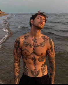 a man with tattoos on his chest standing in the water at the beach looking off into the distance