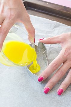 two hands with pink nail polish are pouring yellow liquid into a glass bowl on top of wax paper