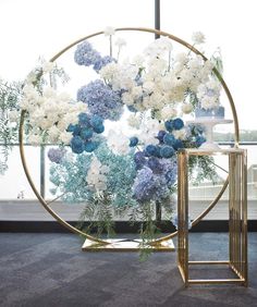 blue and white flowers are arranged in a circular vase on a gold stand next to a window