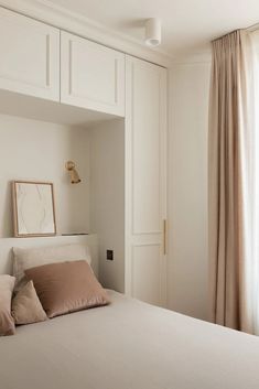 a white bed sitting next to a window in a room with beige curtains and pillows