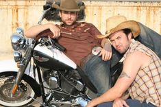 two men sitting on the ground next to a motorcycle with a cowboy hat on it