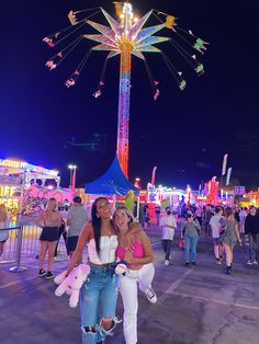 two women pose for a photo in front of a carnival ride at night with other people