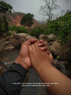 two hands holding each other in front of a river with trees and rocks behind them