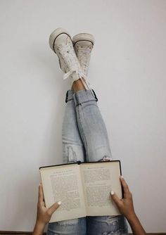 two hands holding an open book in front of the legs of a person wearing sneakers