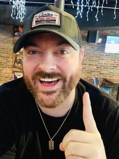 a man with a beard wearing a hat giving the thumbs up sign in front of him