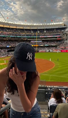 a woman in a yankees hat sitting at a baseball field while talking on her cell phone