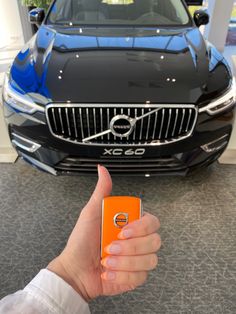 a hand holding an orange cell phone in front of a black volvo car at a showroom